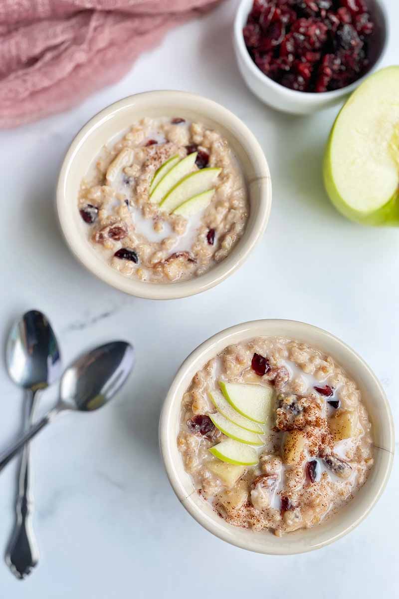 Vertical image of two white bowls filled with a creamy porridge topped with fresh fruit next to a red towel and metal spoons.