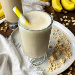 Horizontal image of glasses filled with a creamy beverage, with yellow straws, next to tan powder and fruit.