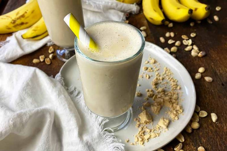Horizontal image of glasses filled with a creamy beverage, with yellow straws, next to tan powder and fruit.