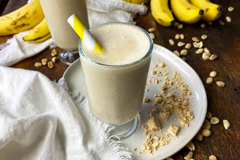 Horizontal image of glasses filled with a creamy beverage, with yellow straws, next to tan powder and fruit.