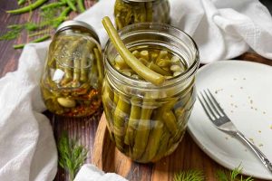 Horizontal image of glass jars filled with skinny vegetables in a brine on a wooden board next to a plate and white towel.