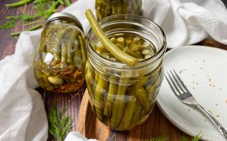 Horizontal image of glass jars filled with skinny vegetables in a brine on a wooden board next to a plate and white towel.