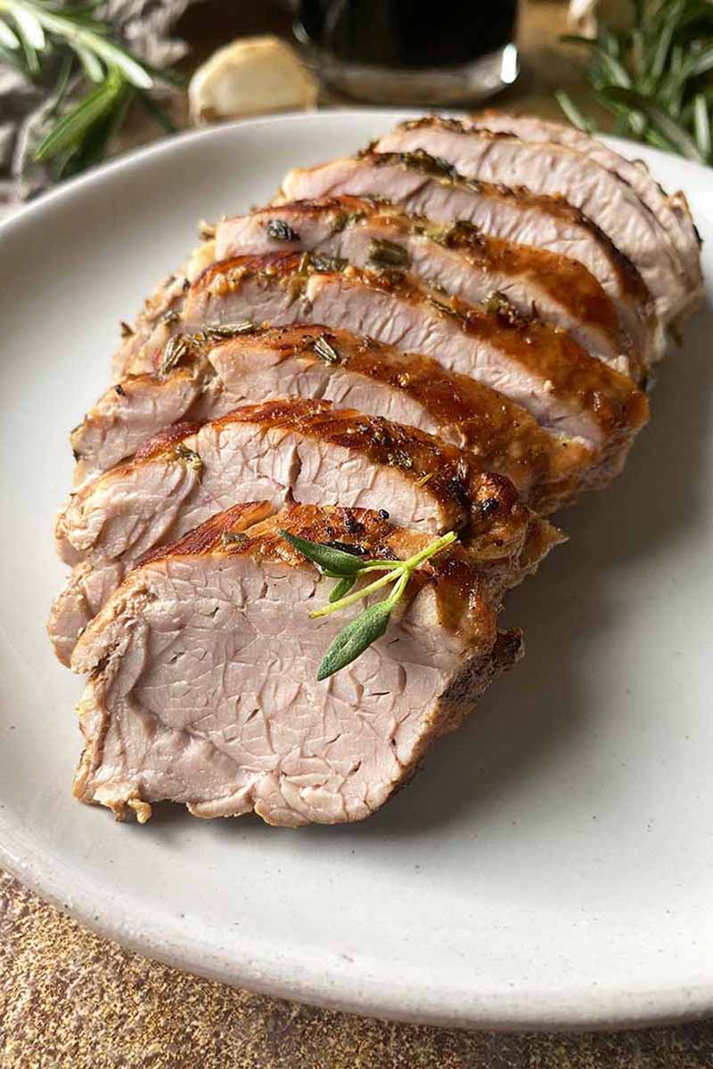 Vertical close-up image of shingled slices of cooked meat with an herb crust on a white plate.