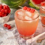 Horizontal image of glasses filled with a light pink beverage filled with ice with a garnish on a pick, next to a white bowl filled with cubes of fresh fruit.
