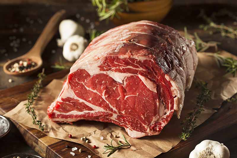 Horizontal image of a large piece of raw beef on paper on a wooden cutting board.