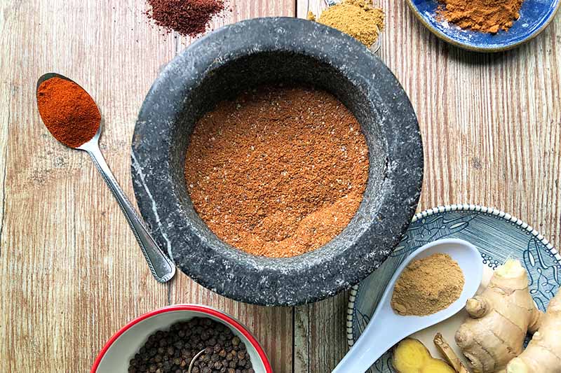 Horizontal image of a gray bowl filled with a deep orange powdery mixture on a wooden table next to spoons with spices, a plate of fresh ginger, and a bowl with whole peppercorns.