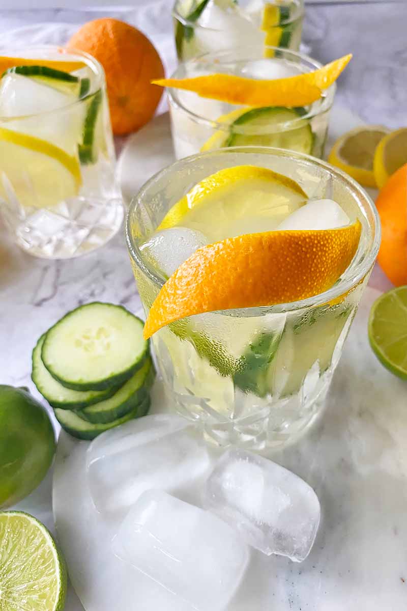 Vertical image of four glasses filled with a beverage, ice cubes, slices of fresh produce, and a piece of rind as garnish.