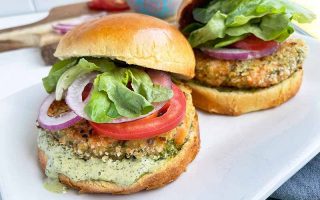 Horizontal image of two sandwiches with seafood patties with an herb sauce, tomatoes, lettuce, and red onions.