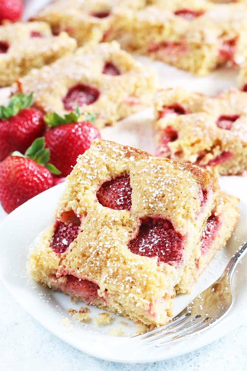 Two slices of strawberry cake are arranged on a white plate with a fork, with more of the dessert and three whole red berries in the background.