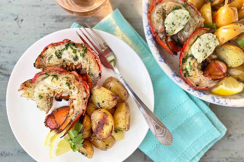 Horizontal image of a small white plate and a large dish with cooked seafood with butter and grilled vegetables next to a blue napkin and glass of wine.