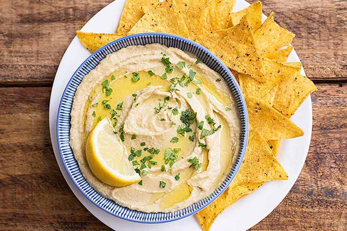 Horizontal top-down image of a creamy bean puree in a bowl garnished with lemon, herbs, and olive oil on a plate next to nachos.
