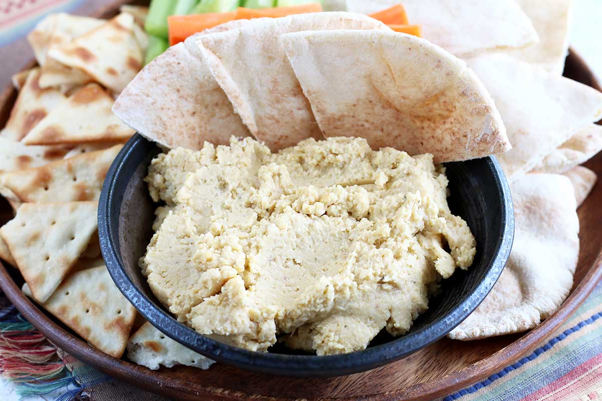 Horizontal image of a thick bean appetizer in a black bowl with pita wedges, chips, and raw vegetables.
