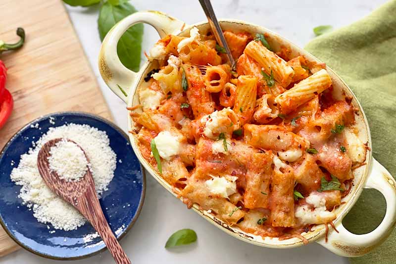 Horizontal image of a green platter with a rigatoni, cheese, and sauce mixture next to a blue bowl with grated cheese.