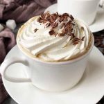 Horizontal image of a white cup filled with a coffee beverage topped with whipped cream and shaved chocolate.