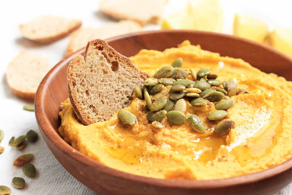 Horizontal image of a wooden bowl filled with a bright orange hummus topped with seeds with a slice of bread inserted into it.