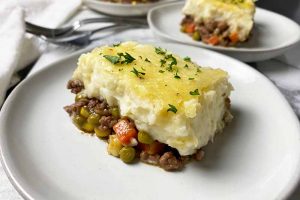 Horizontal image of a large square of a baked casserole topped with parsley on a white plate next to a white towel and metal forks.