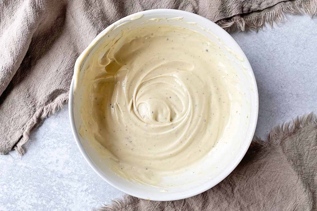 Horizontal image of a creamy dressing in a white bowl next to tan towels.
