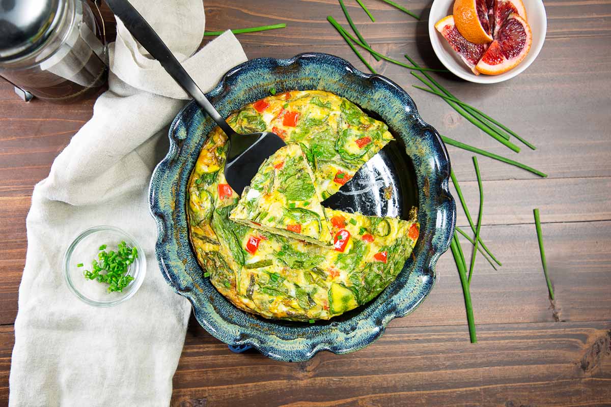 Horizontal top-down image of a breakfast table with coffee, slices of orange in a bowl, and a baked vegetable and egg dish next to a napkin surrounded by scattered fresh chives.