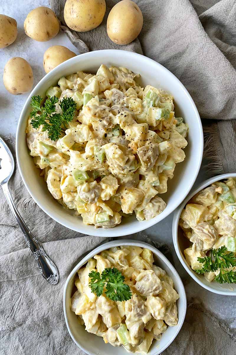 Vertical top-down image of one large bowl and two small bowls filled with potato salad garnished with parsley on tan towels.