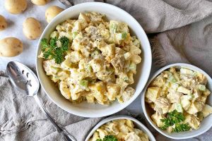 Horizontal top-down image of a large and small bowl of a creamy potato salad garnished with parsley leaves on top of tan towels.