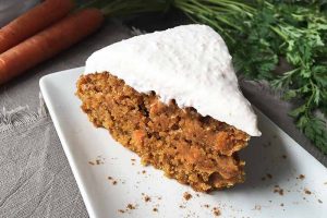 Horizontal image of a slice of a simply decorated orange cake with white icing on a white plate in front of fresh carrots on a tan napkin.