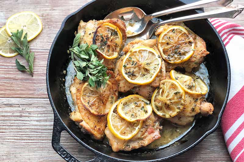 Horizontal image of a pan filled with cooked meat pieces topped with herbs and citrus slices in a sauce next to a red and white towel.