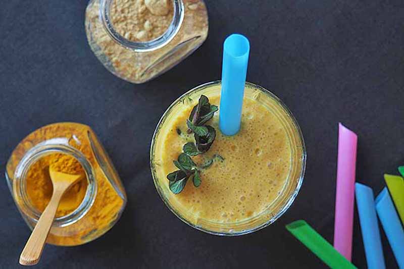 Horizontal top-down image of a glass filled with a creamy orange drink with colorful straws next to jars of ground spices.