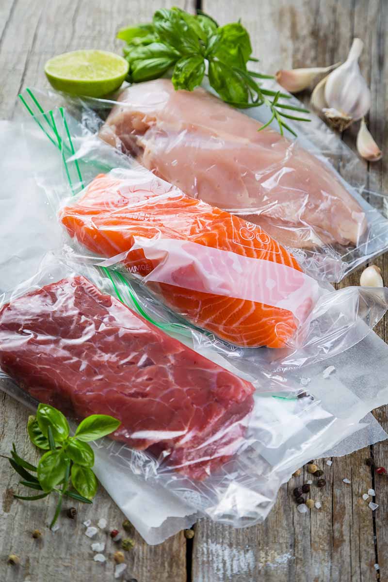 Vertical image of cuts of raw meat in plastic next to herbs.