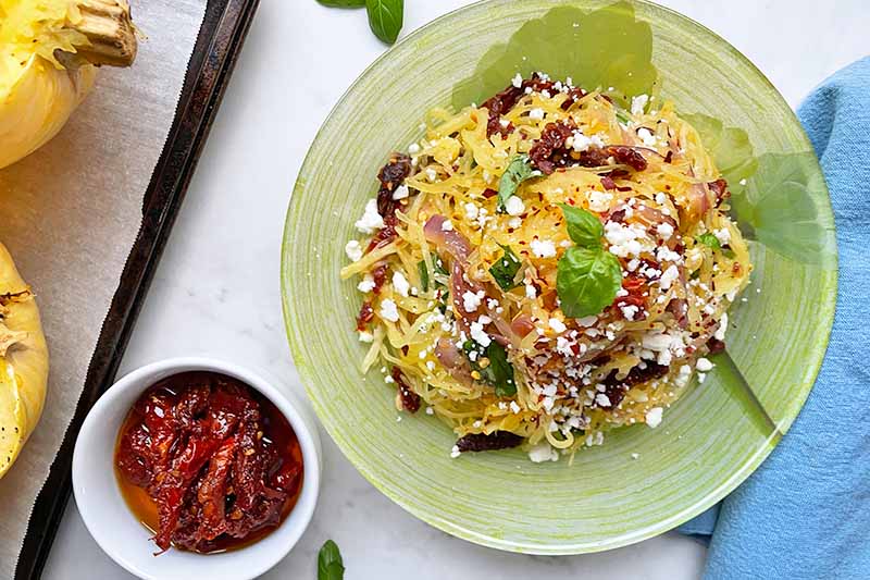 Horizontal image of a green plate filled with strands of yellow vegetables topped with a chunky red sauce with cheese crumbles and basil garnish next to a blue towel and a white ramekin filled with sliced red vegetables.
