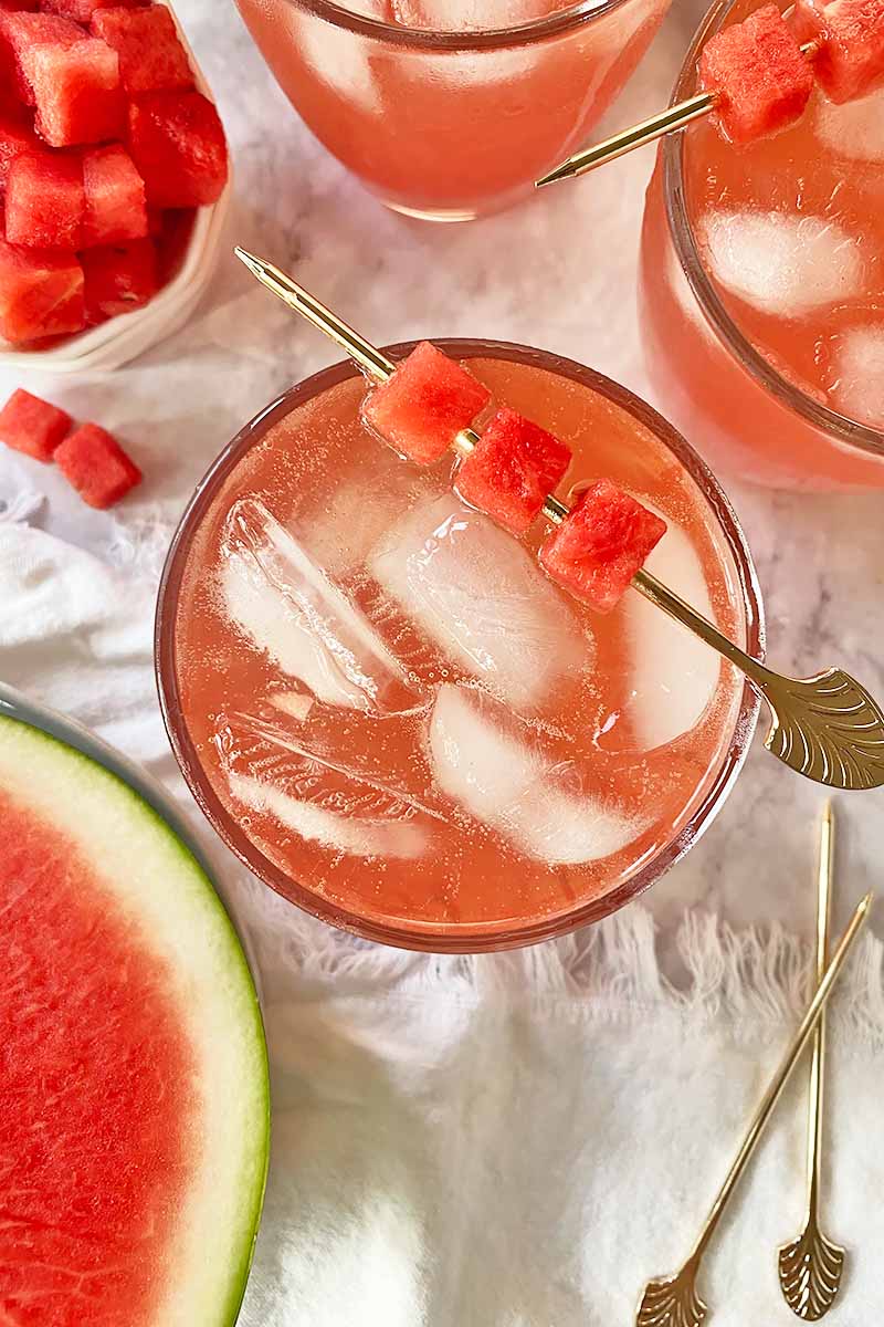 Vertical image of three cups filled with a light pink drink with ice cubes and a garnish with cubes of fruit.