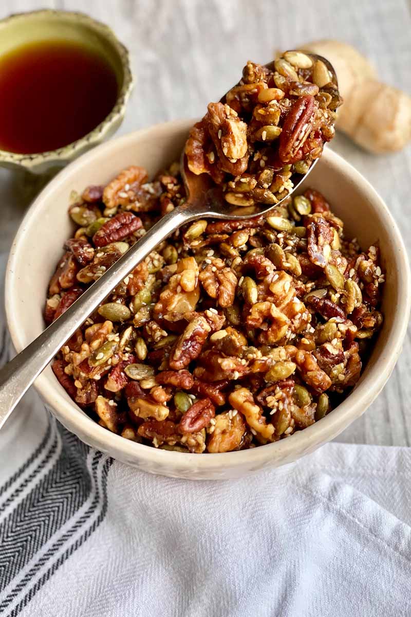 Vertical image of a spoon resting over a white bowl filled with toasted pecans and seeds next to a bowl of maple syrup.