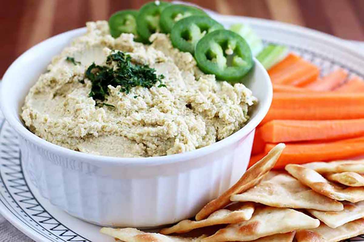Horizontal image of a spicy cilantro and jalapeno pureed bean appetizer in a white ramekin on a plate next to pita chips and raw carrot slices.