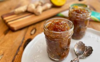 Horizontal image of two jars filled with a dark orange chunky spread on a white plate next to spoons, with ginger in the background on a wooden board.