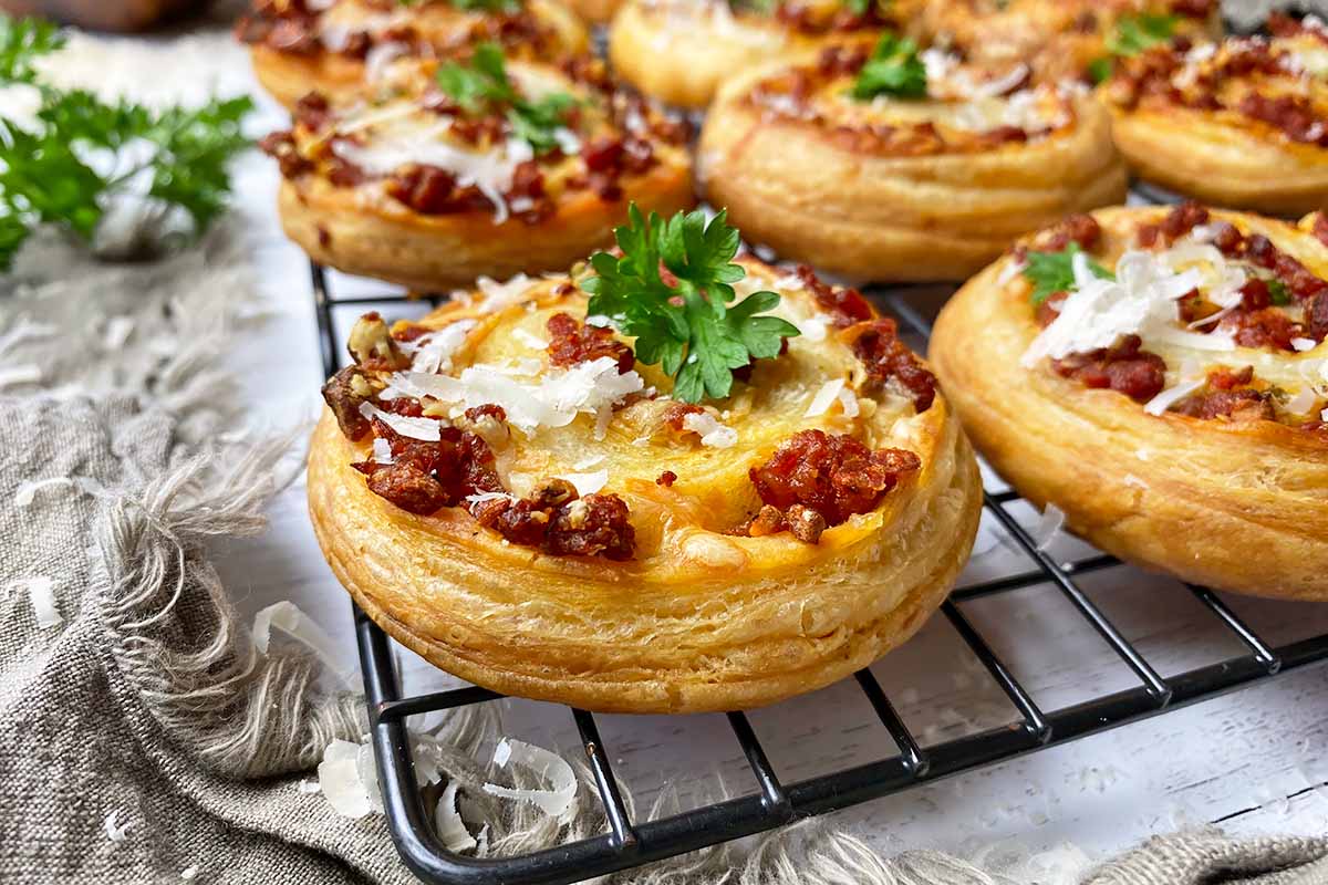 Horizontal image of rows of puff pastry rounds filled with sausage and cheese garnished with parsley on a cooling rack.