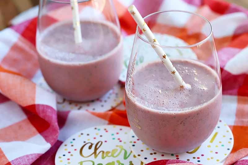 Two stemless wine glasses of a pink fruit smoothie, with paper straws and sitting on cardboard coasters, on a folded and gathered red, orange, and white checkered cloth.