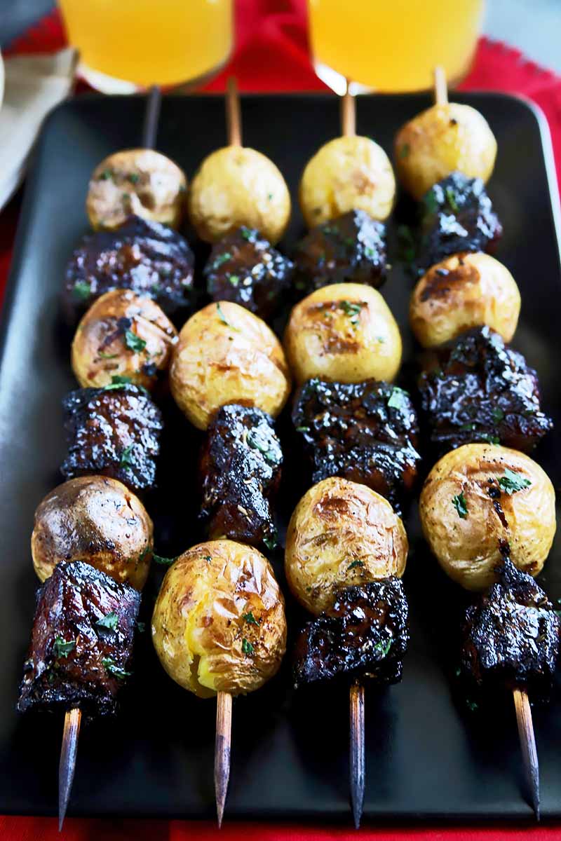 Vertical image of four skewers of meat and potato pieces on a rectangular black plate.