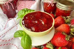 Horizontal image of a bowl of red jelly on a white stand next to basil and fresh fruit.