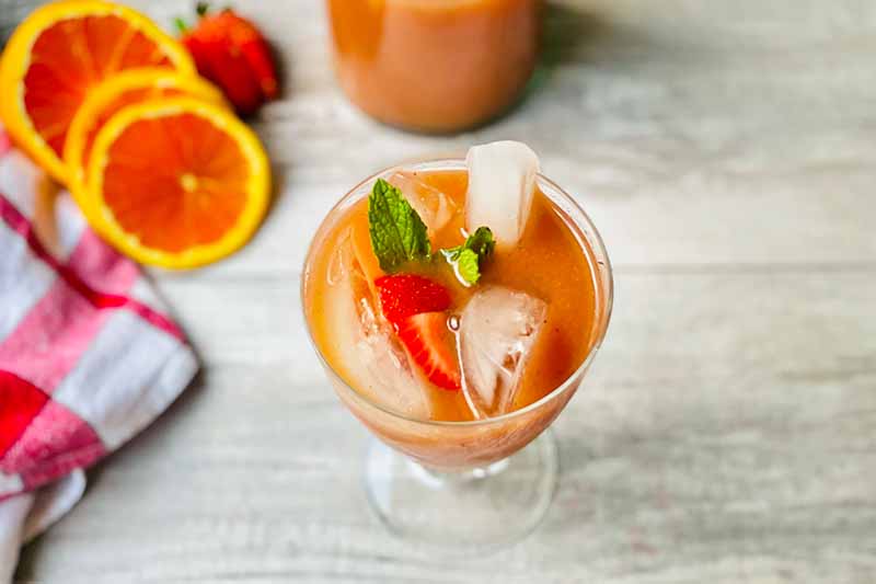 Horizontal image of a drink garnished with ice cubes, fresh fruit slices, and mint leaves.