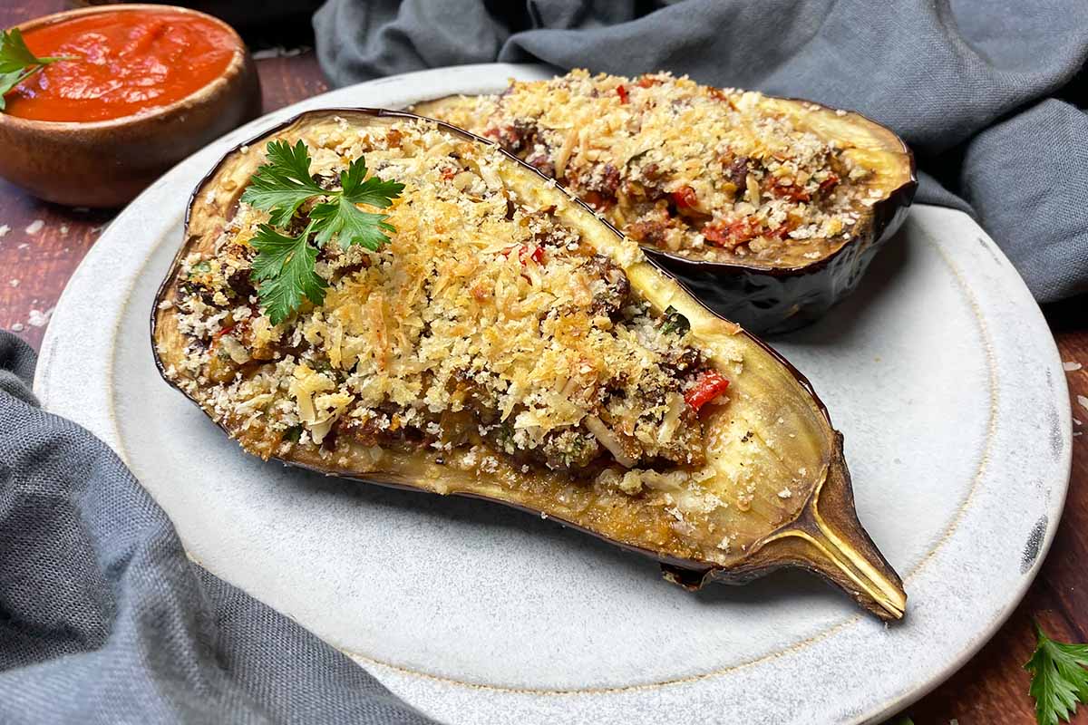 Horizontal image of two eggplant halves on a plate with a meat and mixed veggie filling and breadcrumb topping, next to gray napkins and a bowl of tomato sauce.