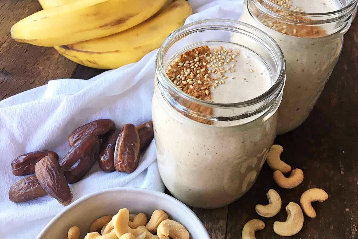 Horizontal image of two glasses filled with a tahini creamy beverage topped with cinnamon and toasted sesame seeds next to dates and cashews.