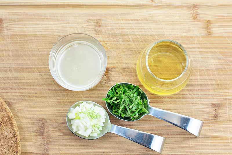 Horizontal image of chopped herbs and fennel in two metal spoons next to a cup of lemon juice and a jar of honey on a wooden surface.