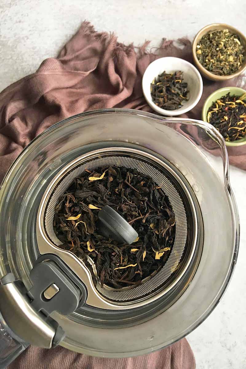 Vertical image of a metal basket with with tea leaves, next to a brown towel and bowls with additional tea.