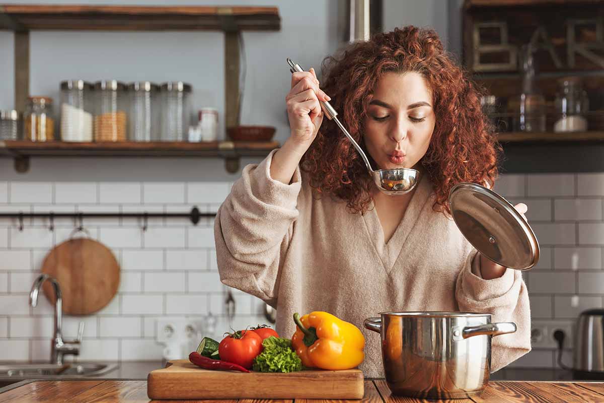 Horizontal image of a woman holding a cooking ladle spoon while testing soup in a pot next to fresh vegetables on a wooden cutting board.