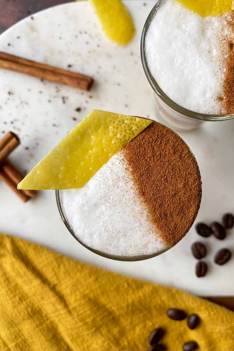 Vertical top-down image of two beverages topped with frothed milk, cinnamon, and lemon rind on a marble counter next to a yellow towel.