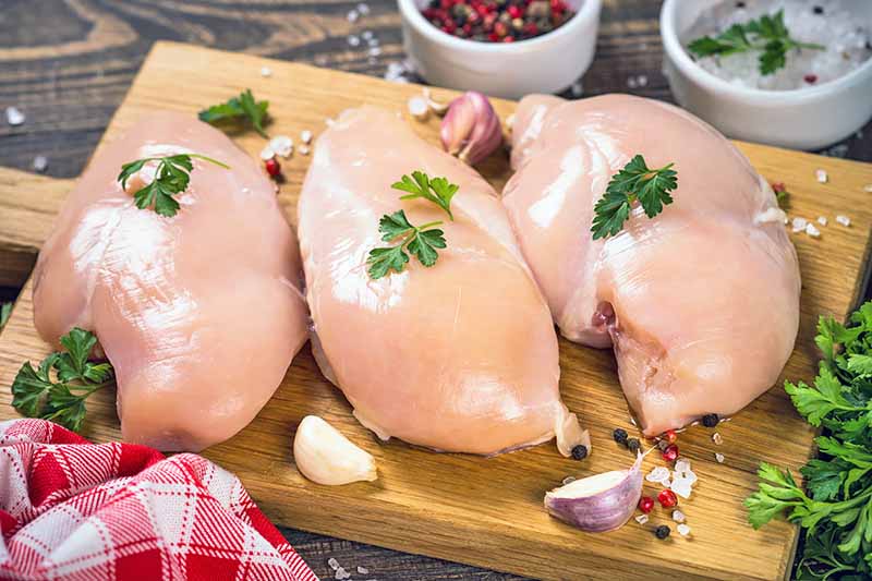 Horizontal image of three raw chicken breasts topped with herbs on a wooden board next to herbs and seasonings and a checkered towel.