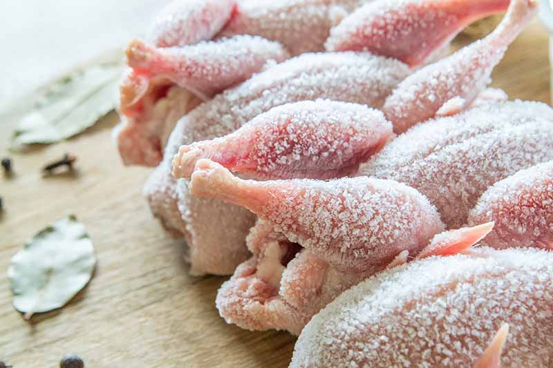 Horizontal closeup image of whole small poultry on a wooden chopping block next to assorted herbs and seasonings.