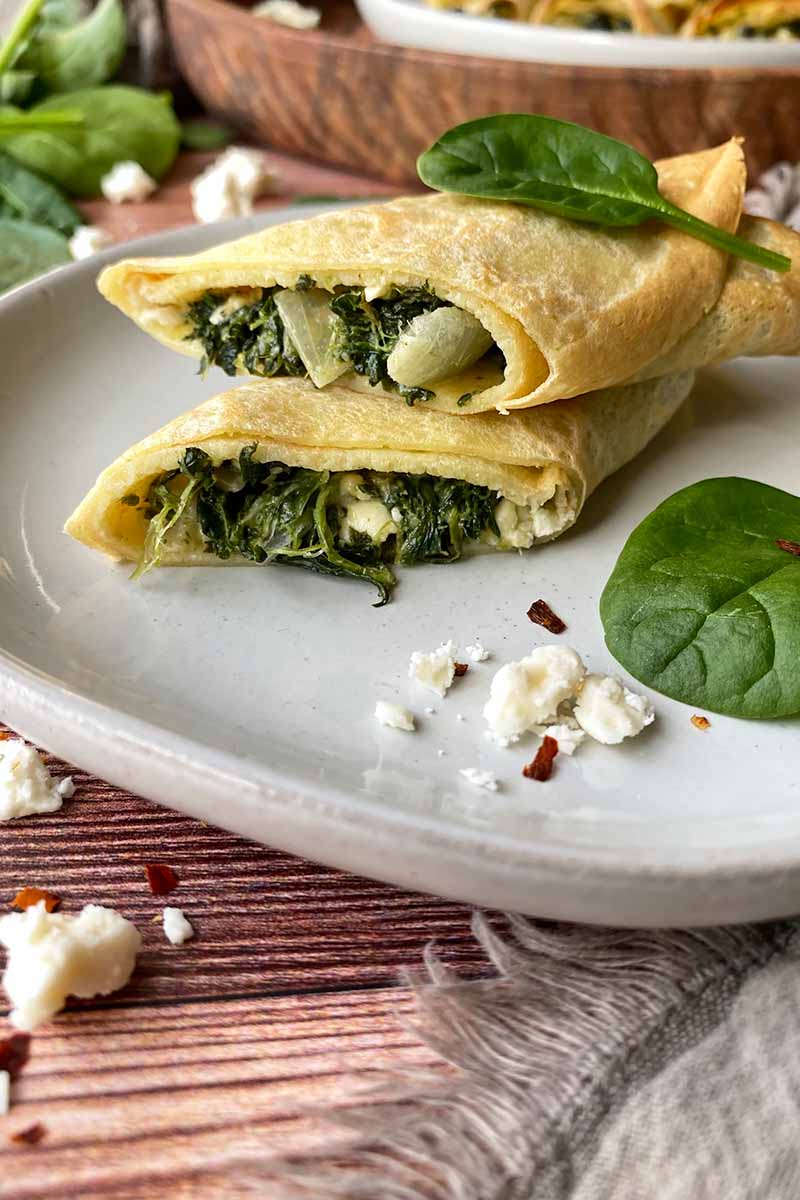 Vertical image of a halved and stack thin pancake with a greens, onion, and cheese stuffing on a plate next to a wooden serving platter and a tan towel.