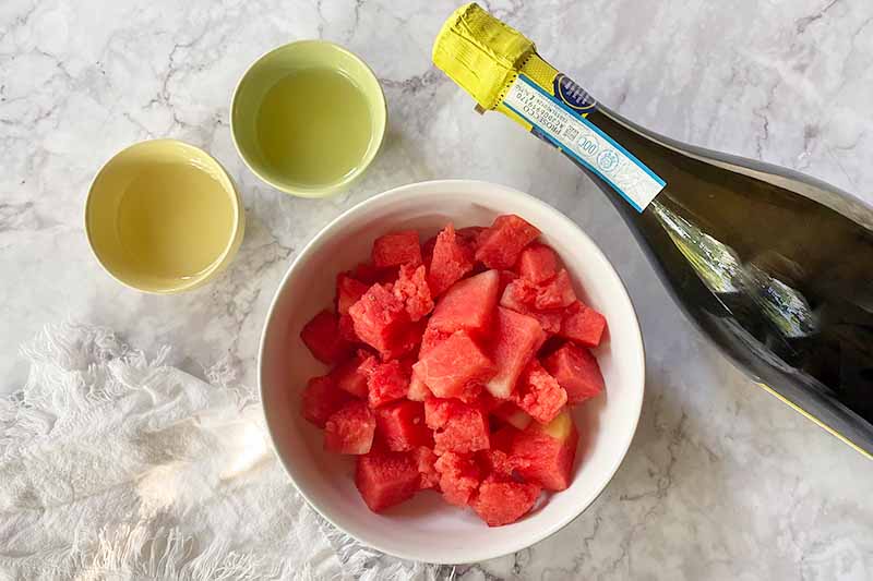 Horizontal image of two small bowls filled with liquids, one large bowl filled with freshly cut fruit, and a bottle of wine.