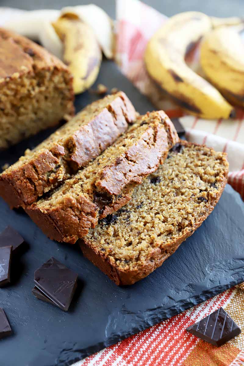 Vertical image of thick slices of a dark yellow baked loaf on a slate next to fruit and chocolate chunks.