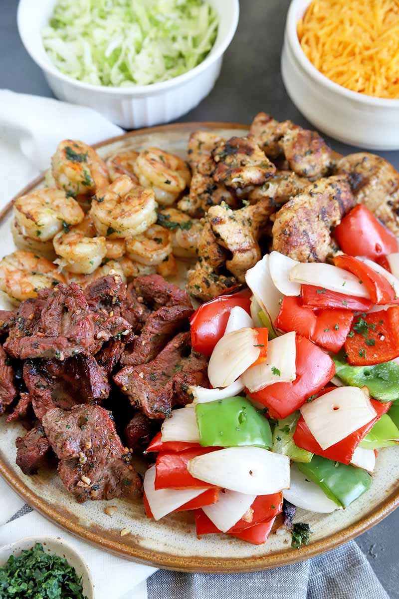 Vertical oblique overhead closely cropped image of a cream-colored ceramic serving platter with a brown rim piled with grilled steak, chicken, shrimp, green and red bell peppers, and onions, sprinkled with green chopped herbs, on a gray surface with small white dishes of cilantro, shredded lettuce, and shredded yellow cheese, on a folded and gathered white cloth on top of a gray surface.